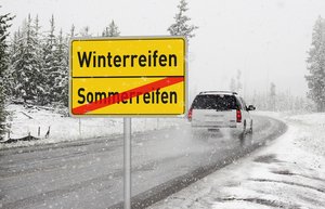 Ortsausgangsschild, oben steht Winterreifen, darunter, durchgestrichen Sommerreifen. Im Hintergrund fährt ein Auto durch eine Schneelandschaft