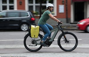 Eine Frau fährt auf einem Pedelec auf einem Radweg. Im Hintergrund sind zwei Parkende Autos zu sehen.