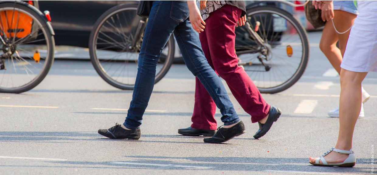 Drei Menschen überqueren eine Straße von rechts nach links. Man sieht nur ihre Beine und Füße.
