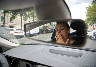 Eine Frau sitzt am Steuer ihres Wagens und gähnt. Die Kameraaufnahme ist durch den Rückspiegel gemacht.