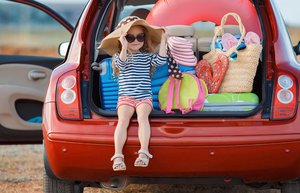 Der Kofferraum eines roten Kleinwagens ist geöffnet. Darin sind Koffer und Taschen für den Urlaub verstaut. Ein kleines Mädchen mit Sonnenhut und Brille sitzt links im Kofferraum und lächelt den Betrachter an. Foto: Adobe Stock