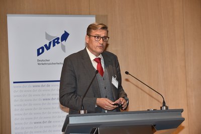 Auf dem Podium hinter dem Rednerpult steht Dr. Michael Weyde. Er trägt einen grauen Anzug, eine rote Krawatte, ein weißes Hemd und hat eine Brille auf. Seine Haare sind kurz geschnitten.