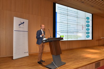 Auf dem Podium hinter dem Rednerpult steht Klaus Böhm. Er trägt eine braune Hose, ein weißes Hemd und ein blaues Sakko. Dazu hat er eine braune Krawatte an.