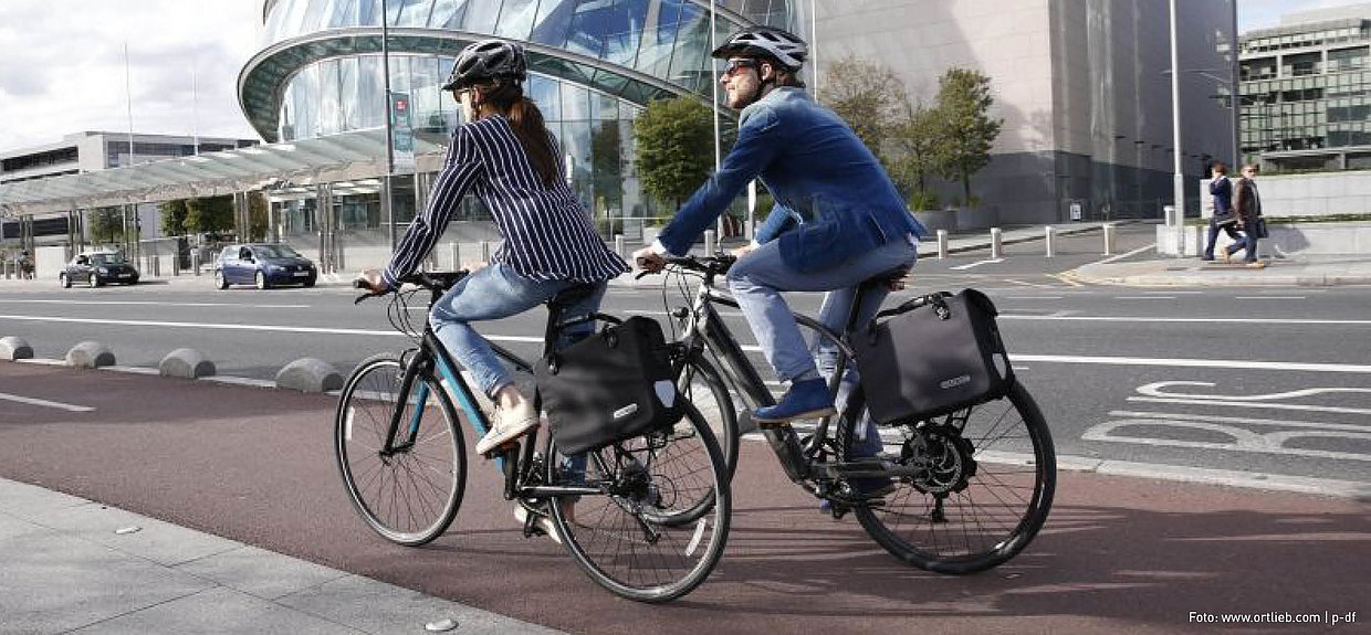 Programm Sicherheit für den Radverkehr. Foto: www.ortlieb.com | p-df Ein Mann und eine Frau in Business-Outfits fahren Fahrrad.
