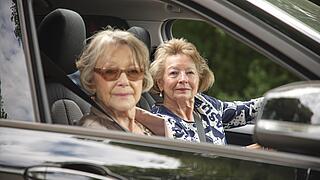 Zwei ältere Damen sitzen in einem schwarzen Auto. Eine von ihnen ist die Fahrerin des Pkws, die andere sitzt auf dem Beifahrersitz. Sie blicken in Richtung der Kamera, die ins Beifahrerfenster hinein fotografiert.