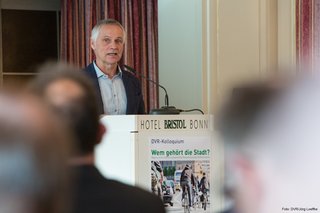 Prof. Dr.-Ing. Jürgen Follmann steht auf dem Podium hinter einem Pult. Die Aufnahme ist aus der Perspektive des auf Stühlen sitzenden Publikums gemacht.