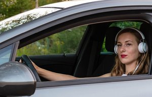 Eine Frau sitzt hinter dem Steuer eines Autos. Sie trägt weiße Kopfhörer auf den Ohren und blickt aus dem Autofenster.