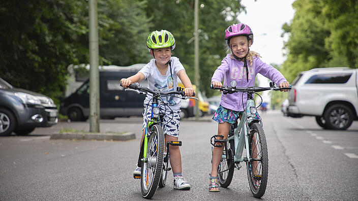 Ein Junge und ein Mädchen im Alter von sechs Jahren stehen an ihren Fahrrädern auf einer wenig befahrenen Straße. Beide tragen einen Helm.