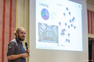 Friedemann Goerl, Fußverkehrsbeauftragter der Stadt Leipzig, steht vor dem Publikum. Er hält ein Mikrofon in der linken Hand. In der rechten Hand hält er einen Präsenter. Hinter ihm an der Wand ist ein Bild projiziert. Es zeigt unter anderem den prozentualen Anteil nach Verkehrsbeteiligungsart in Leipzig aus dem Jahr 2008. 
