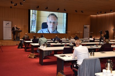 Das Publikum sitzt im Saal mit großen Abständen zwischen den einzelnen Plätzen. Auf der Leinwand ist Dr. Benjamin Krenberger zugeschaltet.