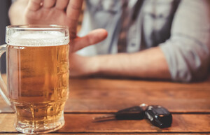 Eine Person sitzt an einem Holztisch und macht ein Handzeichen „Stopp“. Auf dem Tisch steht ein volles Bierglas und neben dem Glas liegt ein Autoschlüssel. 