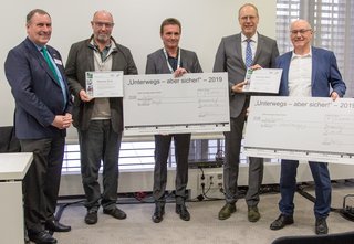 Fünf Männer stehen nebeneinander, zwei halten große Schilder hoch, zwei weitere kleinere Urkunden.