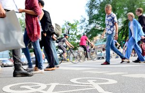Fußgänger und Radfahrer überqueren bei Tageslicht und sommerlichen Temperaturen die Straße. Im Hintergrund warten Autos auf die Weiterfahrt.