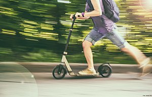 Ein Mann fährt schnell auf einem E-Scooter. Er trägt kurze blaue Hosen, ein lila T-Shirt und einen schwarzen Rücksache. Der Hintergrund ist verschwommen und grün. 