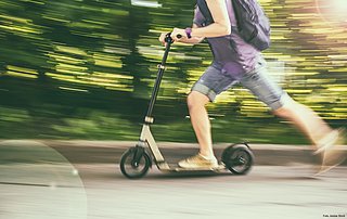 Ein Mann fährt schnell auf einem E-Scooter. Er trägt kurze blaue Hosen, ein lila T-Shirt und einen schwarzen Rücksache. Der Hintergrund ist verschwommen und grün. 