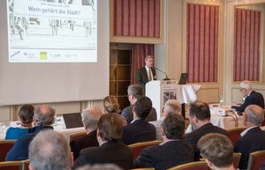DVR-Präsident Prof. Dr. Walter Eichendorf auf dem Podium hinter einem Pult. Die Aufnahme ist aus der Perspektive des auf Stühlen sitzenden Publikums gemacht. Ein an die Wand projiziertes Bild zeigt den Kolloquiumstitel „Wem gehört die Stadt?“