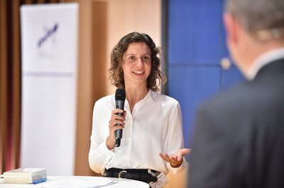 Charlotte Heppner hat lockige braune Haare. Sie trägt eine weiße Bluse und eine schwarze Hose. In ihrer rechten Hand hält sie das Mikrofon. Sie blickt zu Stefan Grieger und damit in die Kamera. Stefan Grieger ist mit dem Rücken zur Kamera im Bild platziert.