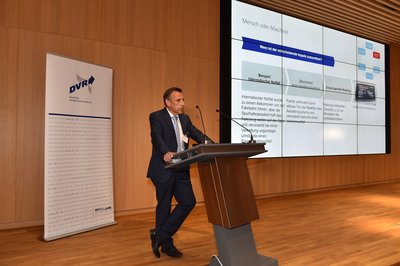 Hinter dem Rednerpult auf dem Podium steht ein Mann. Er trägt einen schwarzen Anzug, eine blaue Krawatte und ein weißes Hemd. 