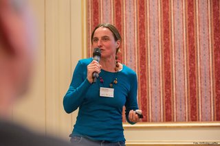 Irene Senfter, Ökoinstitut Südtirol, steht vor dem Publikum. Sie hält das Mikrofon vor ihren Mund und schweift mit dem Blick über das Publikum (was nicht mit abfotografiert ist).