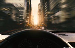 Der Blick geht durch die Windschutzscheibe eines fahrenden Autos. Die Umgebung aus Häusern und Straße ist verschwommen aufgrund der hohen Geschwindigkeit des Autos. Am Horizont geht langsam die Sonne unter.