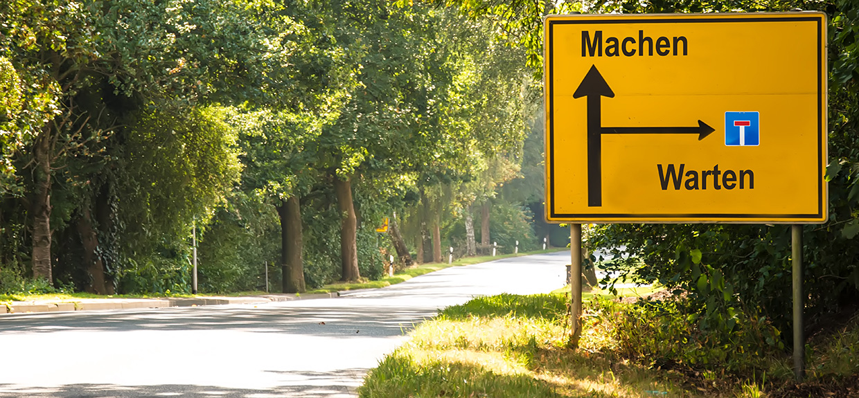Landstraße mit einem fiktiven Straßenschild: Geradeaus steht "Machen", nach Rechts steht eine Sackgasse mit "Warten"
