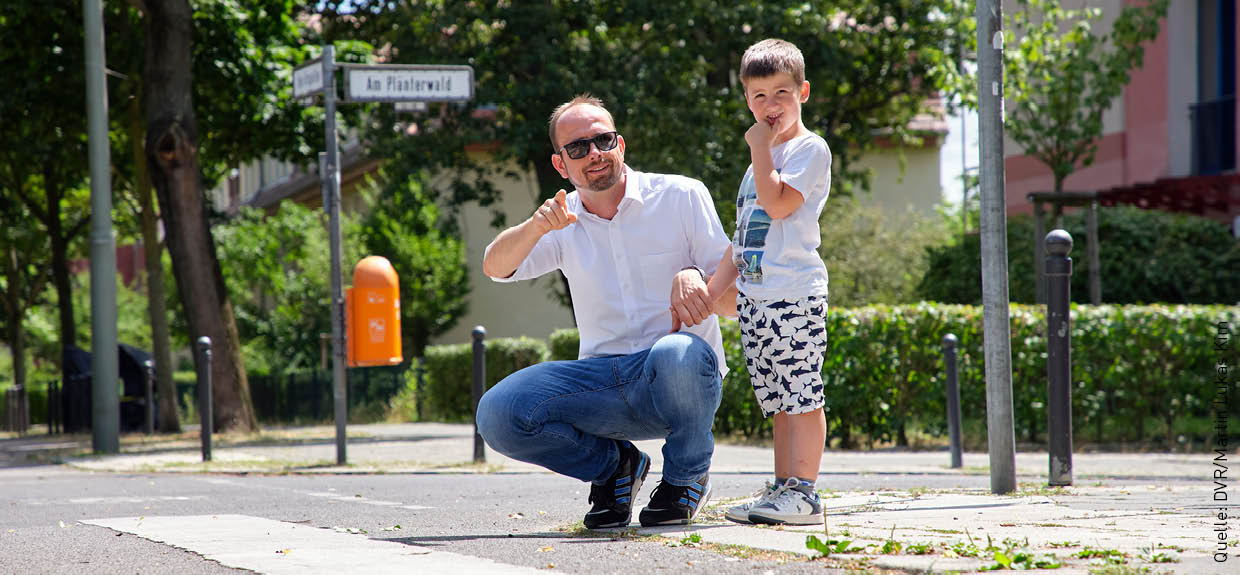 Ein Mann hockt neben einem kleinen Jungen. Er hält die Hand des Kindes. Beide schauen nach rechts in Richtung der Straße, die sie überqueren wollen. Es ist ein sonniger Tag.