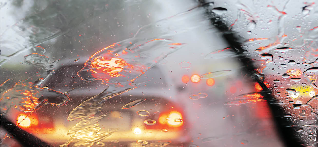 Durch die Windschutzscheibe eines Autos sind die Lichter eines vorausfahrenden Fahrzeugs zu erkennen. Auf der Windschutzscheibe sind Regentropfen, die mit leichten Schlieren von den Scheibenwischern zur Seite geschoben werden.
