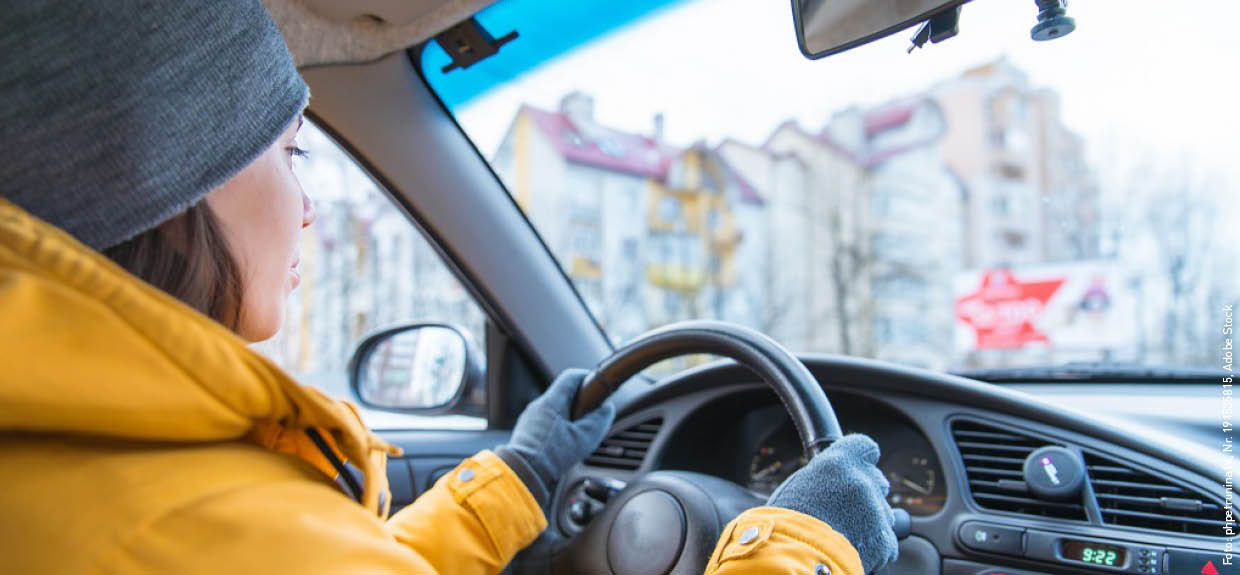 Eine Frau sitzt in einem Auto hinter dem Steuer. Sie trägt Winterkleidung mit einer gelben Daunenjacke, grauen Handschuhen und einer grauen Mütze.